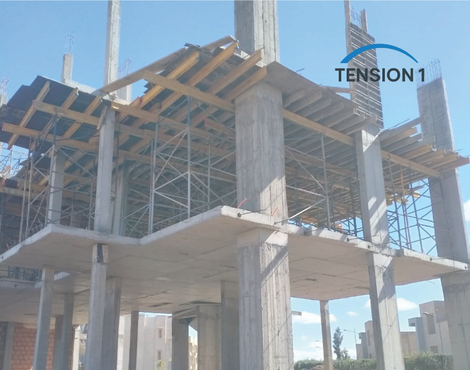 A multi-story concrete residential building under construction with active formwork and extensive metal scaffolding visible, showing preparatory stages for a post-tension slab installation against a blue sky.